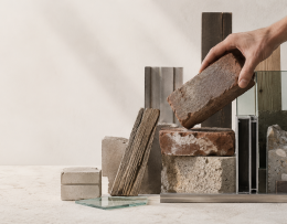 arrangierte wiederverwendete Baustoffe auf neutralem Hintergrund, alte Ziegel, Holzstücke, Glas, Metallprofile und Betonbruch. eine Hand hält prüfend einen alten Ziegel
