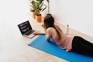 Tipps für mehr Bewegung im Homeoffice. Zu sehen ist eine junge Frau in Sportklamotten, die auf einer Yogamatte liegt und an ihrem Laptop arbeitet.