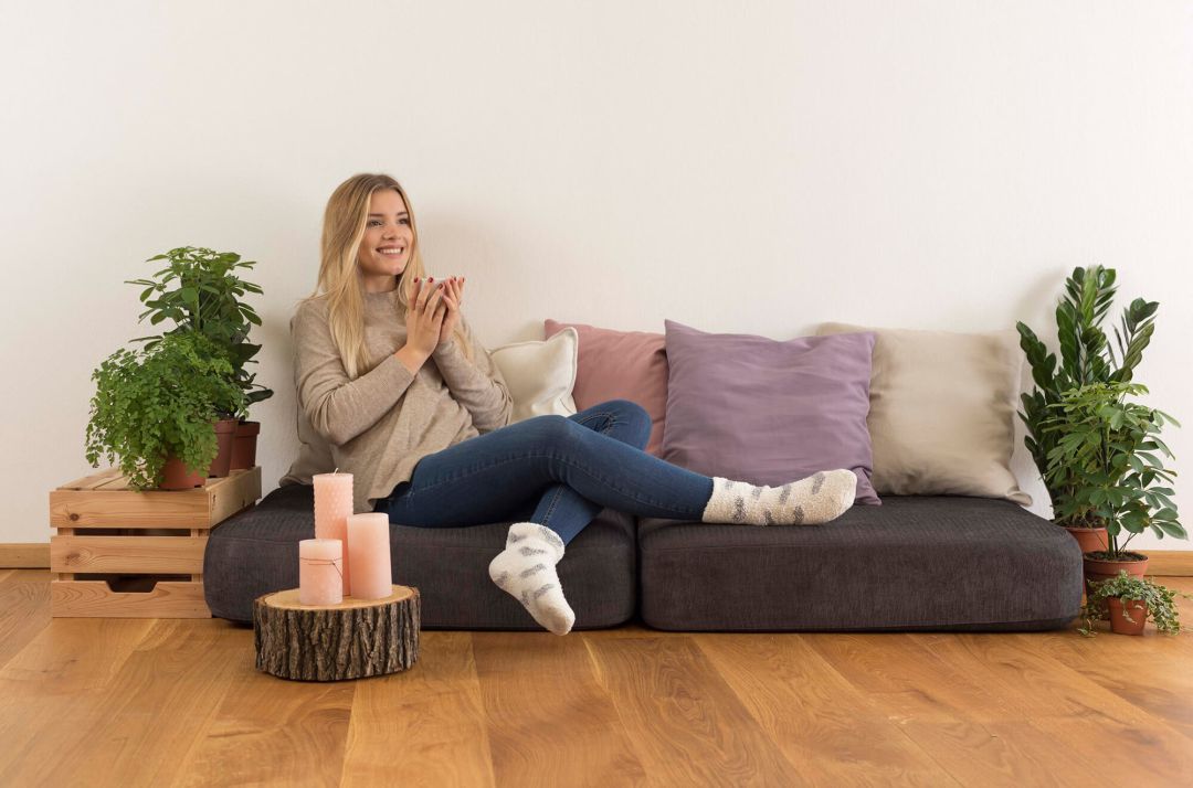Lehmputz im Innenbereich verarbeiten: zu sehen ist eine gemütliche Couch in einem mit Lehmfarbe gestrichenen Wohnzimmer. Eine junge Frau sitzt auf der Couch, dekoriert ist mit Pflanzen und einem Couchtisch aus Holz.