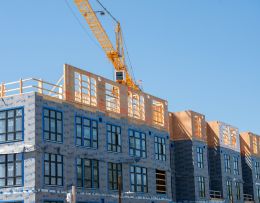Großes Gebäude, das mittels Modulbau errichtet wird, ein Kran hebt gerade ein Fertigteil auf die Baustelle, einige Etagen sind schon fertig.