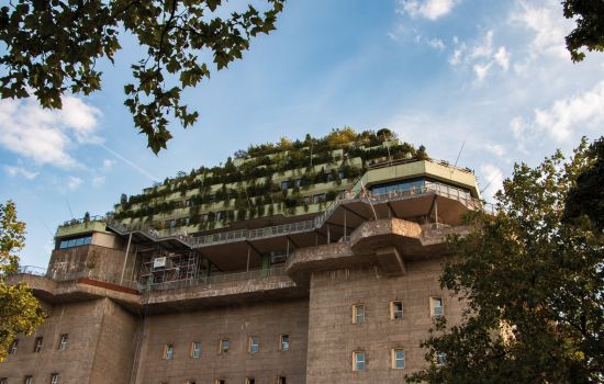 Der begrünte Bunker in Hamburg mit mehreren Etagen, umgeben von Bäumen, vor einem blauen Himmel.