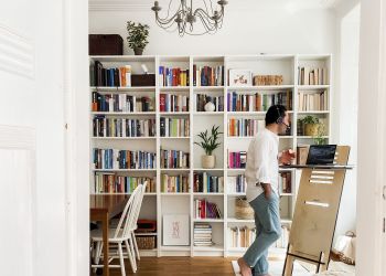 Zu sehen ist ein Wohnzimmer mit großer Regalwand  und einem Esstisch. Ein Mann steht mit einem Headset an einem Stehschreibtisch.