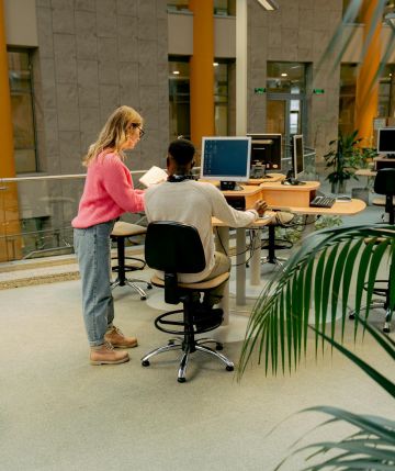 Ein Mann sitzt an einem PC, eine junge Frau steht mit Papieren neben ihm