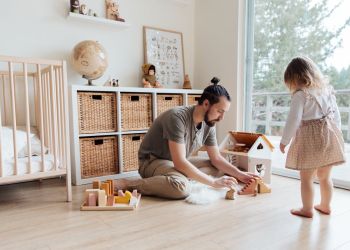 Ein helles, natürlich eingerichtetes Kinderzimmer, in dem ein Mann mit ein Mann mit seiner kleinen Tochter im Puppenhaus spielt.