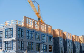 Großes Gebäude, das mittels Modulbau errichtet wird, ein Kran hebt gerade ein Fertigteil auf die Baustelle, einige Etagen sind schon fertig.