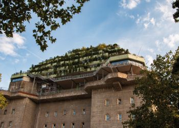 Der begrünte Bunker in Hamburg mit mehreren Etagen, umgeben von Bäumen, vor einem blauen Himmel.