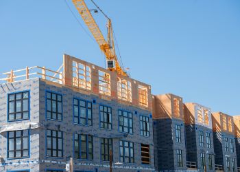 Großes Gebäude, das mittels Modulbau errichtet wird, ein Kran hebt gerade ein Fertigteil auf die Baustelle, einige Etagen sind schon fertig.