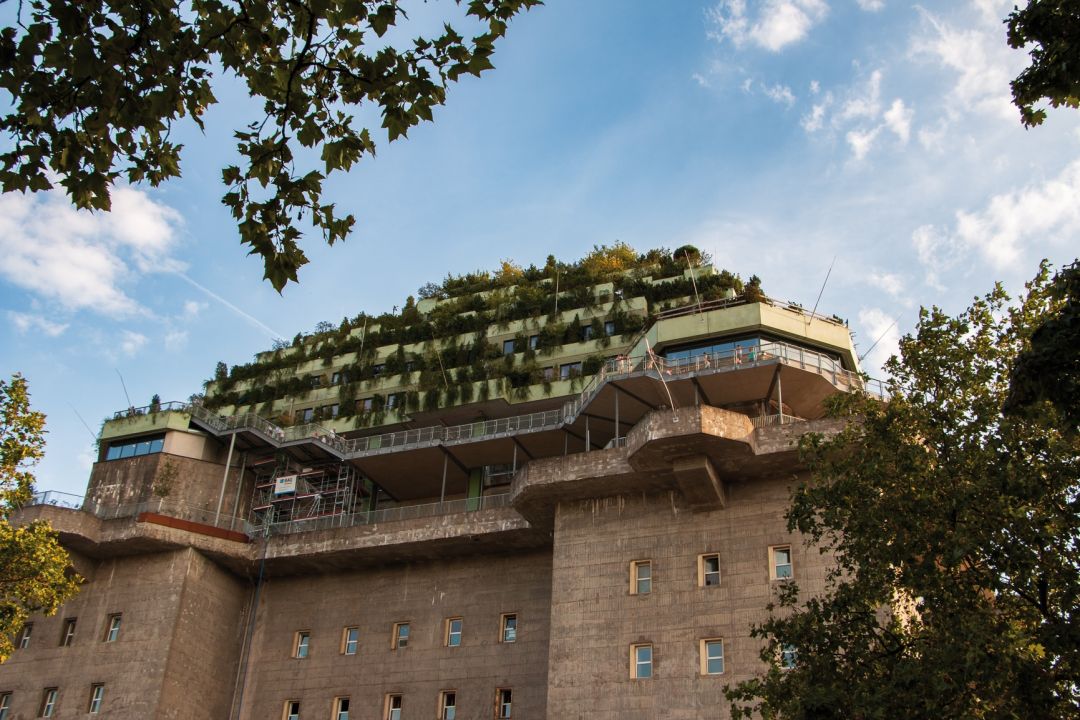 Der begrünte Bunker in Hamburg mit mehreren Etagen, umgeben von Bäumen, vor einem blauen Himmel.