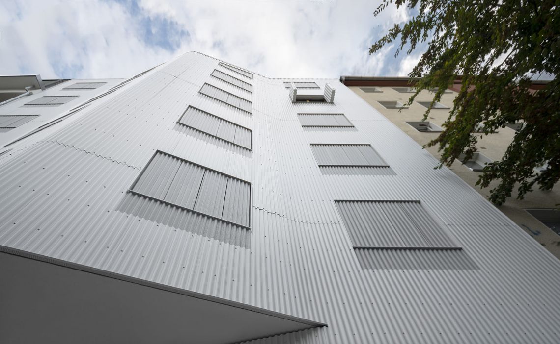 Die symmetrisch und streng gerastert angelegte Straßenfassade in Hybridbauweise ist mit einer nahezu flächenbündigen Außenhaut aus gewelltem Aluminiumblech überzogen.